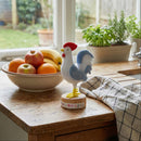 Decorative rooster figurine on a kitchen counter with a bowl of fruit and plants in the background.